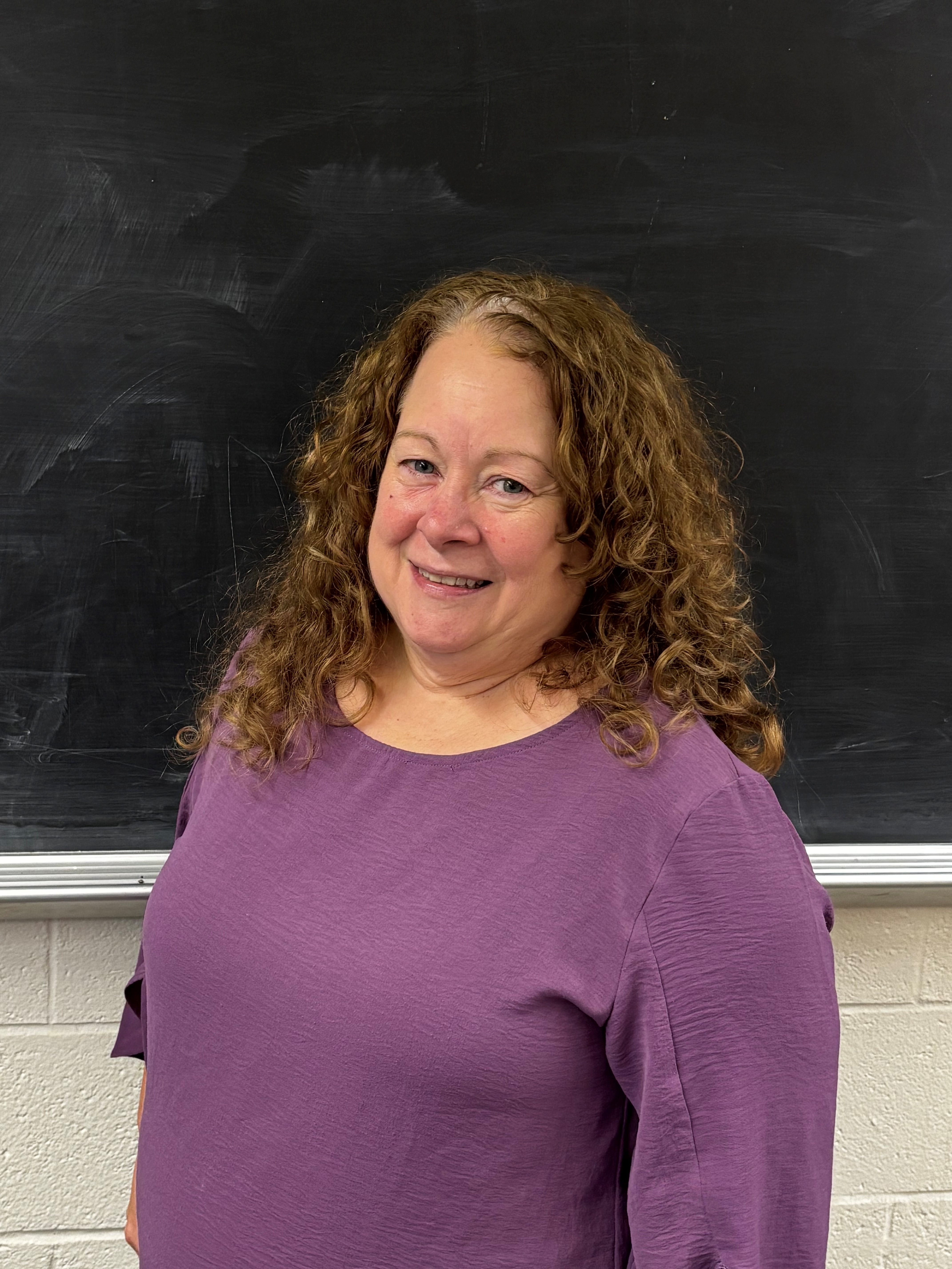 Debbie Webb Portrait of a woman with long curly hair smiling in front of a blakcboard, wearing a purple shirt